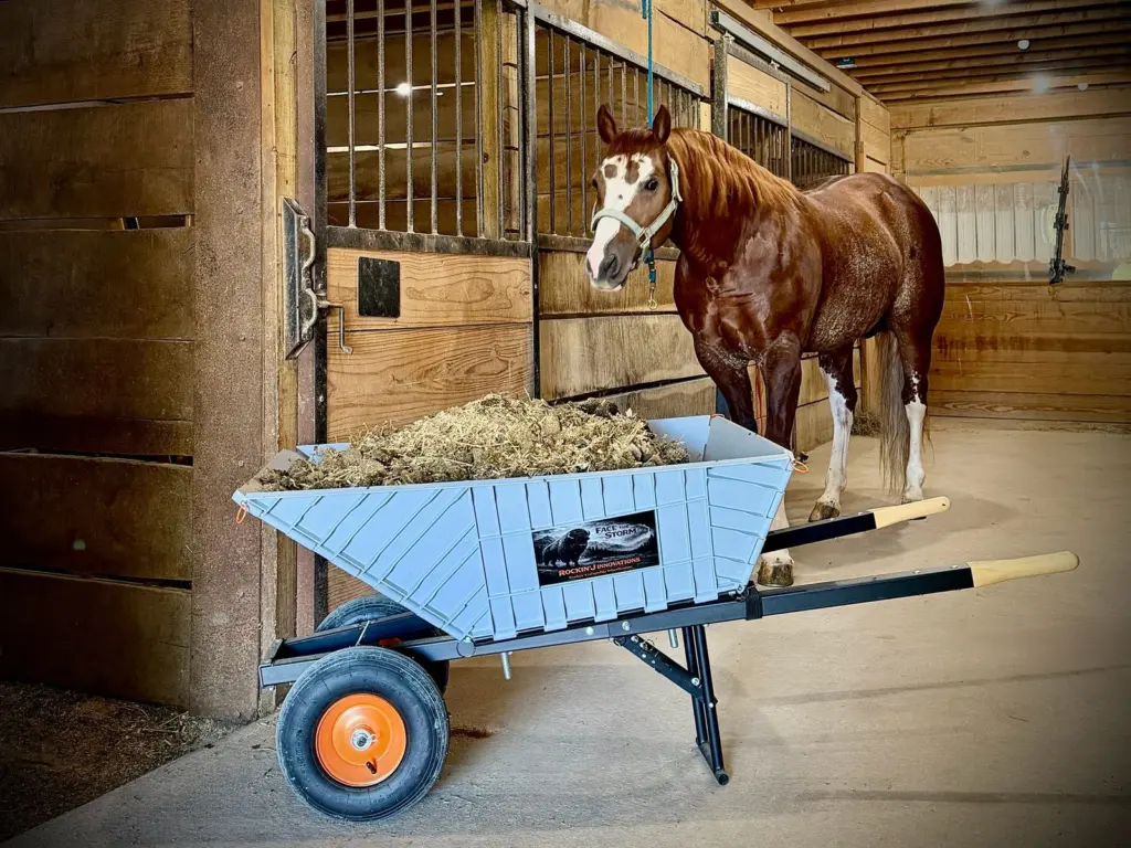 The Workin' Collapsible Wheelbarrow loaded with hay in a barn aisle, a horse looking on from behind it